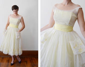 Puede incluir: Vestido vintage hasta la rodilla en un tono amarillo suave. El vestido presenta una capa transparente, un corpiño ajustado y una faja amarilla ancha en la cintura. Delicados bordados florales adornan los bolsillos y el dobladillo.