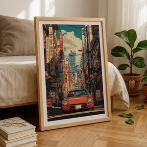 May include: A framed vertical art print featuring a red sports car driving down a narrow city street lined with tall buildings, utility poles, and Japanese signage. The illustration uses a vibrant colour palette with deep shadows and bright highlights.