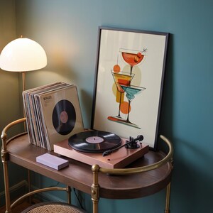 May include: A framed art print featuring three stacked cocktail glasses in red, orange, and blue tones, displayed on a wooden surface next to a copper-colored record player, a stack of vinyl records, and a white lamp with a dome-shaped shade.