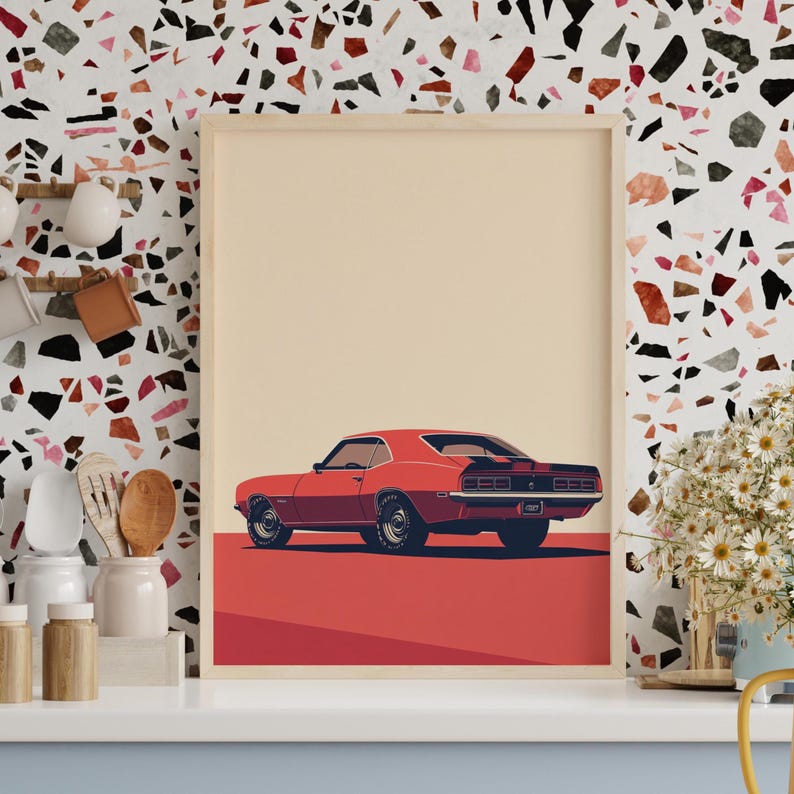May include: A framed art print featuring a stylized illustration of a red muscle car viewed from the rear three-quarter angle. The car has black racing stripes, dark wheels, and a chrome bumper, set against a solid cream background and red base.
