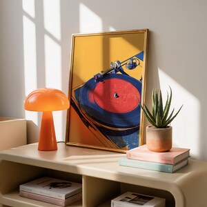 May include: A framed art print featuring a stylized illustration of a record player with a red vinyl disc, positioned next to an orange mushroom-shaped lamp, a potted aloe vera plant, and two stacked books with light-colored covers.