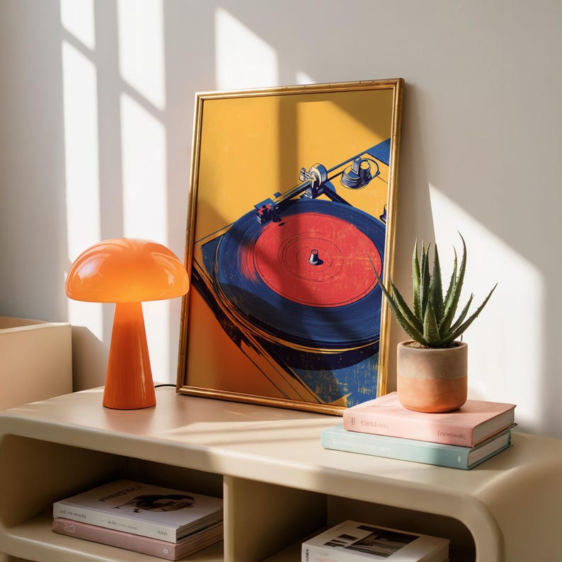May include: A framed art print featuring a stylized illustration of a record player with a red vinyl disc, positioned next to an orange mushroom-shaped lamp, a potted aloe vera plant, and two stacked books with light-colored covers.