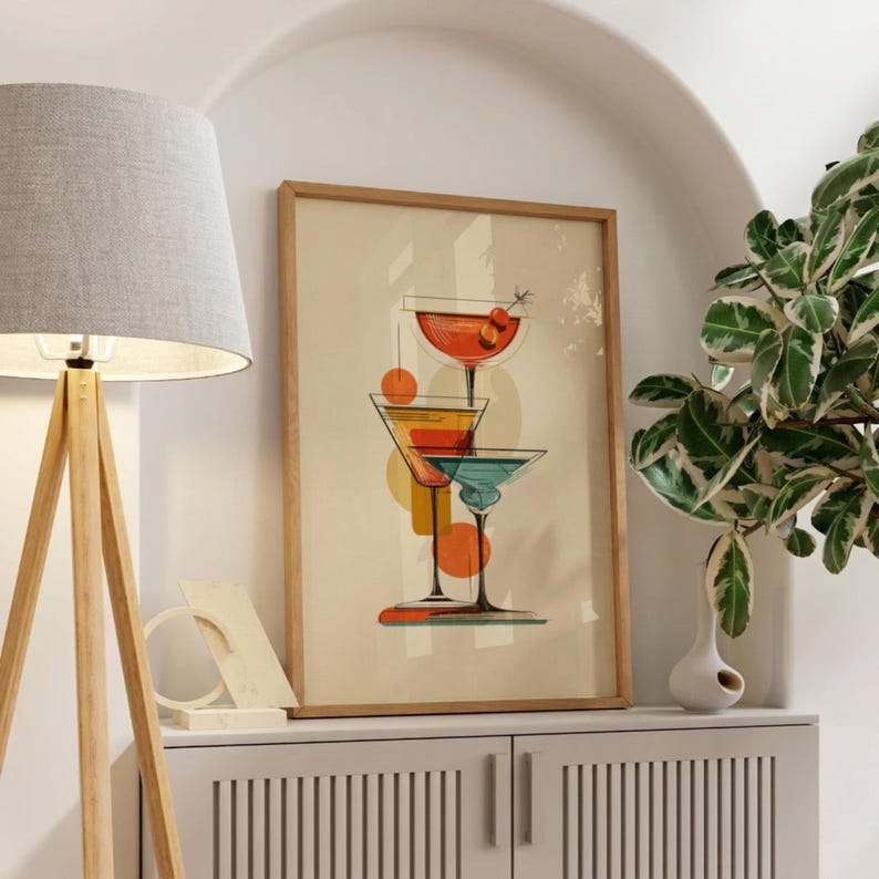 May include: A framed wall art print featuring a vertical stack of three cocktail glasses in shades of orange, yellow, and teal against a cream background. The illustration includes abstract geometric shapes and a small cherry garnish on the top glass.