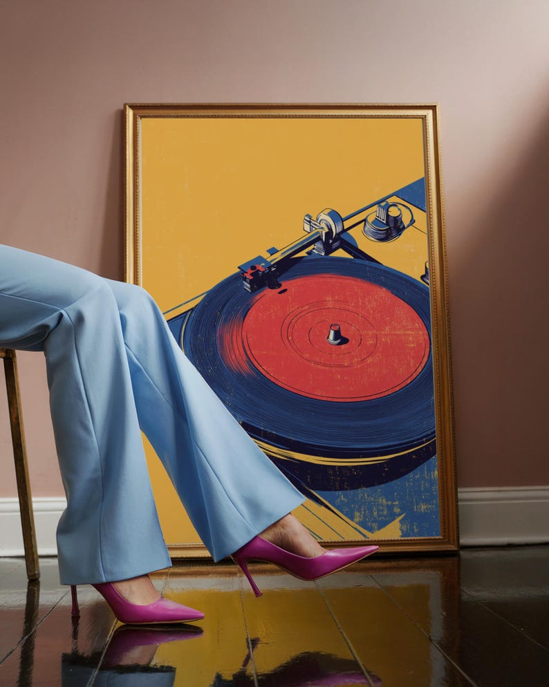 May include: A framed rectangular wall art print featuring a stylized graphic illustration of a record player with a red vinyl record. The artwork uses a color palette of mustard yellow, deep blue, and red, encased in a thin gold-toned frame.