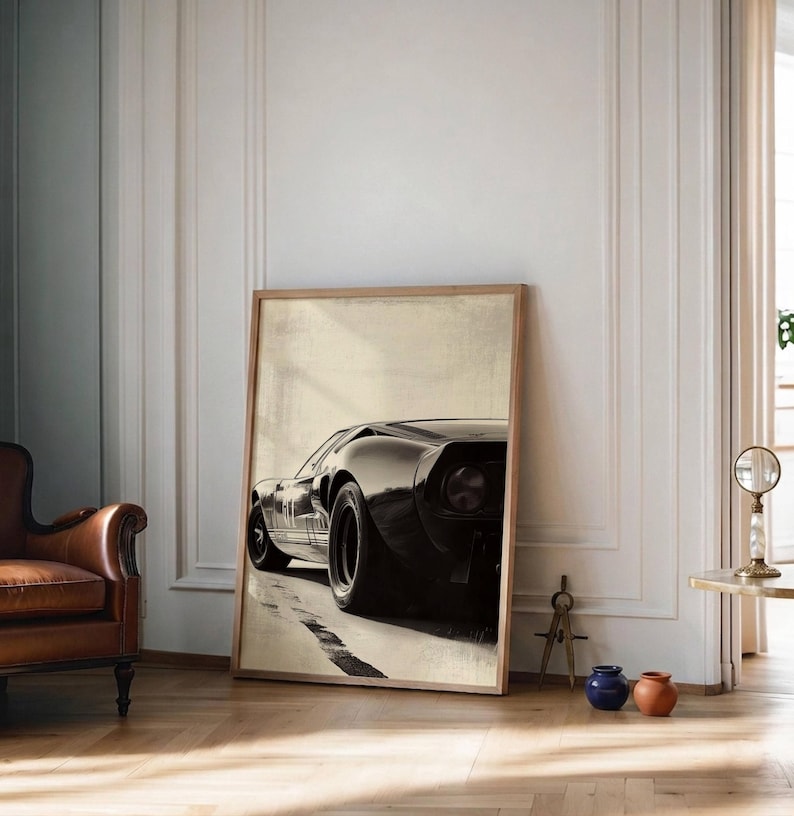 Peut inclure: Une impression encadr&eacute;e d'une voiture de sport noire vintage, vue de l'arri&egrave;re, l&eacute;g&egrave;rement de c&ocirc;t&eacute;. La voiture est plac&eacute;e sur un fond neutre avec une route de couleur claire. L'impression est dans un cadre en bois et est appuy&eacute;e contre un mur.