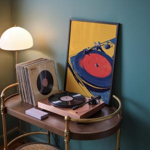 May include: A copper-colored turntable with a black vinyl record sits on a wooden cart next to a stack of vinyl records in sleeves and a framed art print featuring a stylized illustration of a turntable with a red and blue record on yellow.