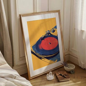 May include: A framed art print featuring a stylized illustration of a record player. The artwork displays a vibrant yellow background with a dark blue turntable and a bright red vinyl record. The frame is metallic gold with a white mat border.