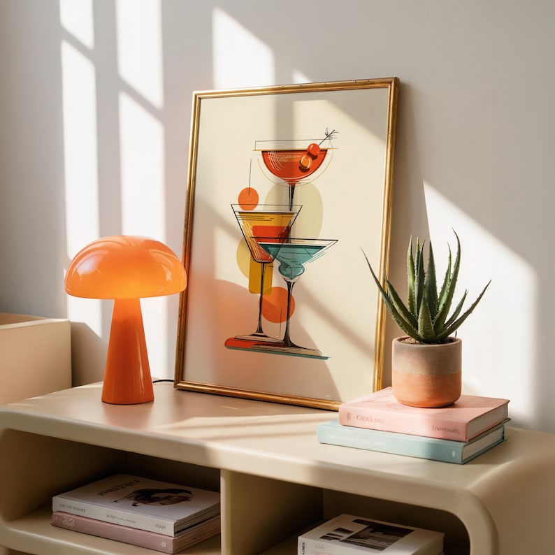 May include: A framed wall art print featuring a stylized illustration of three stacked cocktail glasses in red, yellow, and teal colors. The artwork is displayed in a thin gold frame, showcasing clean black outlines and geometric shapes on a cream background.