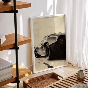 Peut inclure: Impression d'art encadr&eacute;e repr&eacute;sentant une voiture de sport noire vintage. La voiture est vue d'en bas, mettant en valeur son design &eacute;l&eacute;gant et ses gros pneus. Le fond est beige neutre et l'impression est dans un cadre blanc. L'&oelig;uvre est expos&eacute;e sur un sol en bois.
