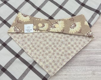 May include: A pet bandana featuring a tan background with a brown and cream floral pattern on the top section and a smaller cream and brown daisy print on the bottom section. It includes two gold-coloured metal snaps and a white label reading Waterwise Dogs.