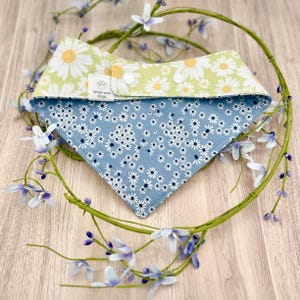 May include: A reversible pet bandana featuring a light green side with a white daisy pattern and a blue side with a small white floral print. The bandana includes white snap closures and a white fabric tag labeled Waterwise Dogs.