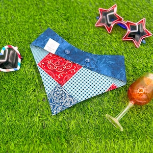 May include: A patchwork dog bandana with red paisley, blue floral, and gingham patterns. The bandana has a blue collar with two button closures. Sunglasses and a wine glass are in the background.