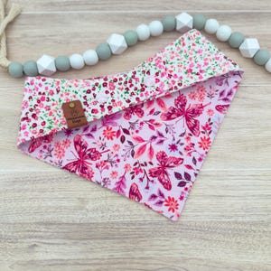 May include: A reversible dog bandana with a floral and butterfly pattern. One side features a pink and red floral print, while the other showcases a pink butterfly design. The bandana includes a brown leather tag with the text "Waterwise Dogs".