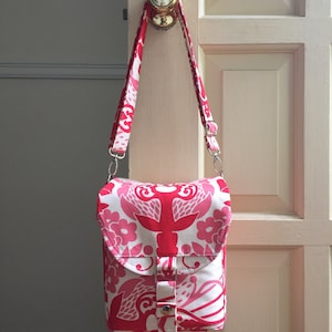 May include: A pink and white floral patterned crossbody bag with a flap closure and a silver buckle. The bag has a long adjustable strap.