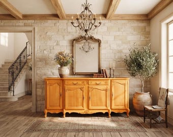 Könnte beinhalten: Ein helles Holz-Sideboard mit vier Schranktüren und zwei mittigen Schubladen vor einer Steinwand. Darüber befinden sich ein rechteckiger Spiegel mit Goldrahmen, ein Kristallleuchter, eine Keramikvase mit Trockenblumen und einige Bücher.