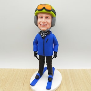 May include: A figurine of a skier wearing a yellow helmet with orange goggles and a blue jacket. The figurine is standing on a white oval base with blue skis. The skier is holding black ski poles.