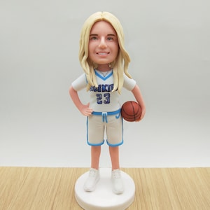 Könnte beinhalten: Figur einer Basketballspielerin mit blonden Haaren, die ein weißes Trikot mit "HUSKIE 23" in Blau trägt. Sie hält einen braunen Basketball und steht auf einer weißen Basis. Die Shorts haben blaue Paspeln.