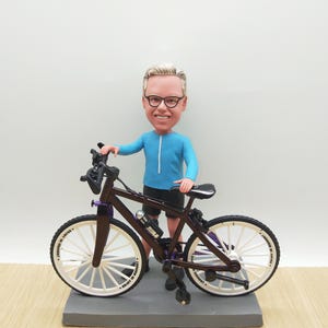 Könnte beinhalten: Figur einer Person mit Brille und blauem Oberteil, neben einem dunkelbraunen Fahrrad mit weißen Rädern. Das Fahrrad hat lila Akzente und eine Wasserflasche. Die Figur steht auf einem grauen Sockel.