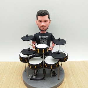 Può includere: Statuetta di un batterista che suona una batteria. Il batterista indossa una maglietta nera con la scritta "Pink Floyd". La batteria ha tamburi neri con dettagli gialli e piatti neri. La statuetta è su una base grigia.