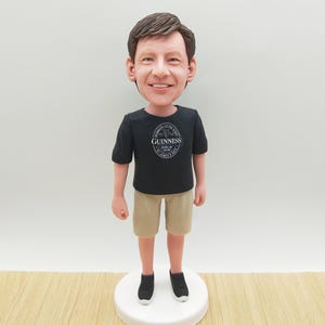 Könnte beinhalten: Figur einer lächelnden Person, die ein schwarzes T-Shirt mit Guinness-Logo, khakifarbene Shorts und schwarze Turnschuhe trägt. Die Figur steht auf einer weißen, kreisförmigen Basis, mit einer hellen Holzmaserung im Vordergrund.
