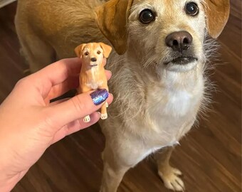 May include: A tan-colored dog stands next to a miniature figurine of a dog. The figurine is held in a person's hand. The dog has brown eyes and a light brown coat. The figurine is a smaller version of the dog.