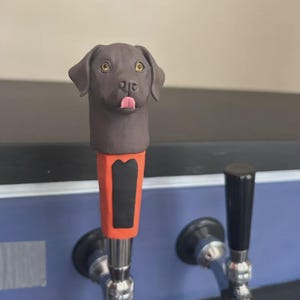 May include: A brown dog-shaped tap handle with a red and black accent. The dog has a pink tongue sticking out. The tap handle is attached to a silver faucet. The background is a blurred blue and black.