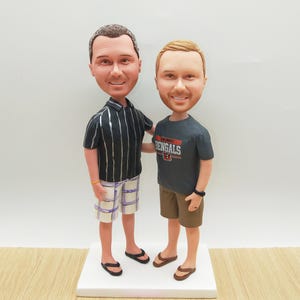 May include: A custom bobblehead figurine featuring two men standing together. One wears a black and white striped shirt and plaid shorts, the other a gray Bengals t-shirt and brown shorts. Both are smiling and wearing sandals, standing on a white base.