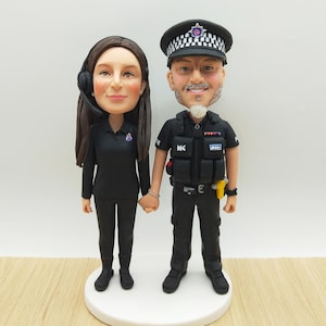 May include: A custom cake topper featuring a couple, a woman in a black uniform with a headset and a man in a police uniform. The figures are holding hands and standing on a white base. The man is wearing a police hat.