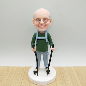May include: A figurine of a smiling, elderly man wearing glasses, a green shirt, light blue pants, and a backpack. He holds two black walking sticks and stands on a white base. The figurine is a cake topper.