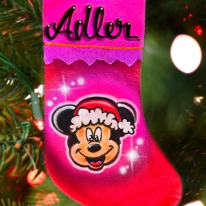 May include: Pink Christmas stocking with a Mickey Mouse illustration and the name "Adler" written in black cursive on the top.