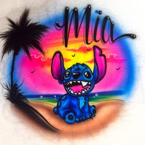 May include: A vibrant illustration featuring a blue cartoon character with large ears and a wide grin, set against a colorful sunset backdrop. The word "Mia" is written above the character in a stylized script. A palm tree and birds are also included.