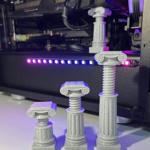 May include: Four white, 3D printed, miniature columns with a fluted design and a decorative capital. The columns are arranged in a row from largest to smallest.