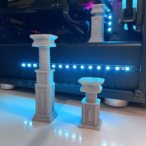 May include: Three light grey 3D printed objects, each featuring a square base, a central threaded screw-like column, and a flat square top. The objects are arranged with two in the foreground and one in the background, displaying a matte finish.