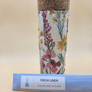 May include: A white ceramic diffuser bottle with a cork lid and a floral design. The bottle is surrounded by a set of reeds and a label that reads "FRESH LINEN SCENTED REED DIFFUSER".