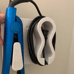 Puede incluir: Una plancha de pelo azul y un soporte de pared blanco moteado para el cable y el enchufe. La plancha tiene un diseño texturizado y un logotipo. El soporte tiene un diseño curvo para sujetar el cable y el enchufe.