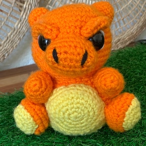 May include: A handmade orange crochet creature with black eyes and a yellow belly and feet. The plush toy is sitting on green grass. The creature has an angry expression.