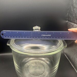 May include: A clear glass casserole dish with a matching lid and side handles. A blue Fiskars ruler is positioned across the top, showing the lid has a diameter of approximately 8.5 inches. The glass is transparent and smooth.