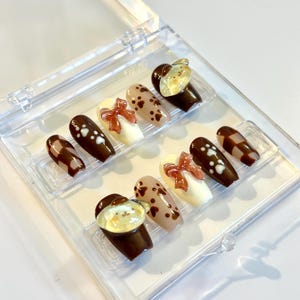 May include: A set of ten press-on nails in a clear plastic case, featuring a brown and cream color palette. Designs include brown and cream checkered patterns, solid brown with white paw prints, cream with 3D brown bows, and brown with bear faces.