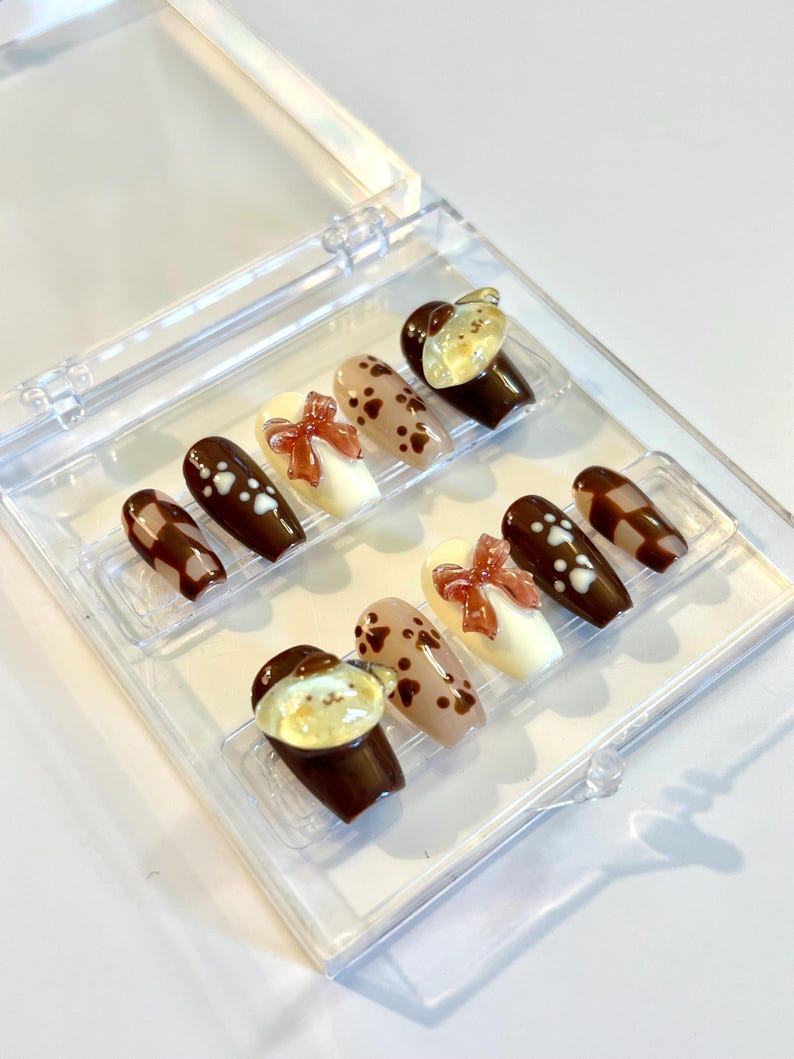 May include: A set of ten press-on nails in a clear plastic case, featuring a brown and cream color palette. Designs include brown and cream checkered patterns, solid brown with white paw prints, cream with 3D brown bows, and brown with bear faces.