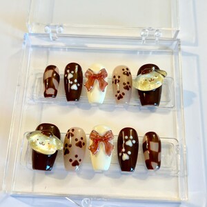 May include: A set of ten press-on nails in a clear plastic case, featuring a brown and cream color palette. Designs include brown and cream checkered patterns, white paw prints on brown, cream bases with 3D brown bows, and yellow dog charms.