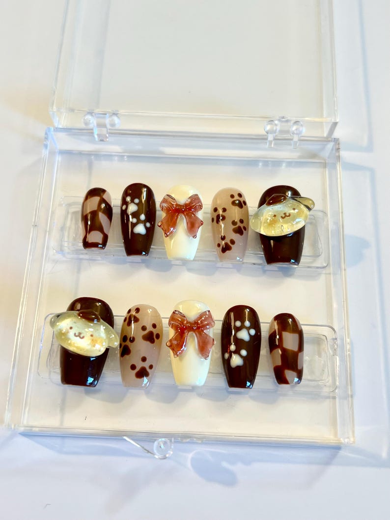 May include: A set of ten press-on nails in a clear plastic case, featuring a brown and cream color palette. Designs include brown and cream checkered patterns, white paw prints on brown, cream bases with 3D brown bows, and yellow dog charms.