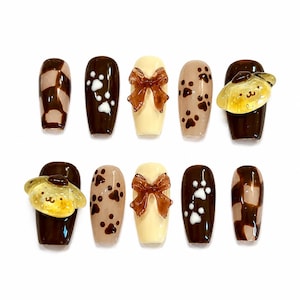 May include: A set of ten press-on nails featuring a brown and cream color palette. Designs include brown and cream checkered patterns, white and brown paw prints, solid cream nails with three-dimensional brown bows, and yellow character charms.