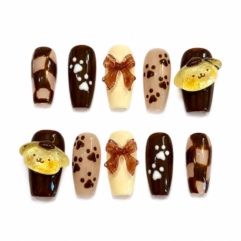 May include: A set of ten press-on nails featuring a brown and cream color palette. Designs include brown and cream checkered patterns, white and brown paw prints, solid cream nails with three-dimensional brown bows, and yellow character charms.