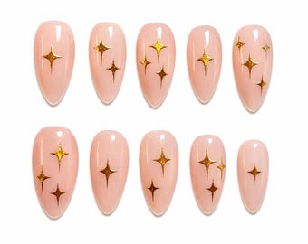 Pode incluir: Um conjunto de dez unhas postiças em formato de amêndoa com uma base rosa pálido. Cada unha tem um acabamento brilhante e é decorada com desenhos de estrelas douradas de quatro pontas de vários tamanhos sobre a superfície.