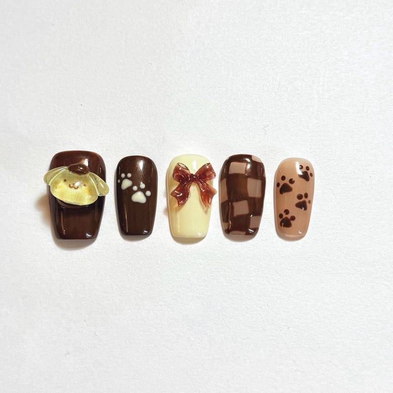 May include: A set of five press-on nails featuring a brown and cream color palette. Designs include a yellow dog character charm, white paw prints on brown, a translucent brown bow on cream, a brown checkered pattern, and brown paw prints on beige.