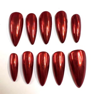 Peut inclure: Dix faux ongles rouges, de forme stiletto et à finition métallique, disposés sur deux rangées sur une surface blanche. Les ongles varient en taille, les plus grands étant en haut et les plus petits en bas. La couleur est un rouge profond et brillant.
