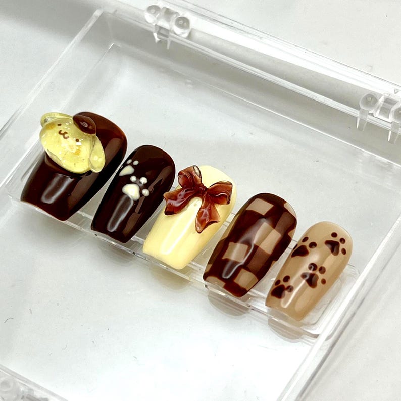 May include: A set of five press-on nails featuring a brown and cream color palette. The designs include a yellow character charm, white paw prints, a brown 3D bow, a brown and cream checkered pattern, and brown paw prints on a light tan base.