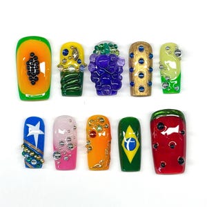 May include: A set of ten press-on nails featuring diverse designs including a papaya motif, a grape cluster, a Brazilian flag, a watermelon slice, and various abstract patterns with metallic beads, rhinestones, and colorful glossy finishes.