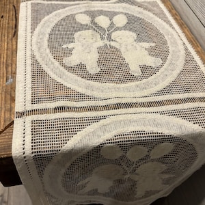 May include: White crocheted table runner featuring a repeating design of two figures holding balloons. The figures are enclosed in a circle and the runner has a grid-like pattern.