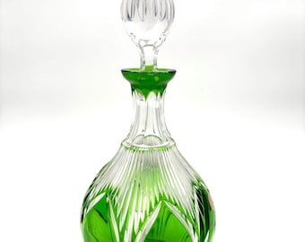 May include: A clear glass decanter with a green interior and a clear, rounded stopper. The decanter has a bulbous body with vertical cuts and a green base. The stopper is also clear and has a ribbed design.